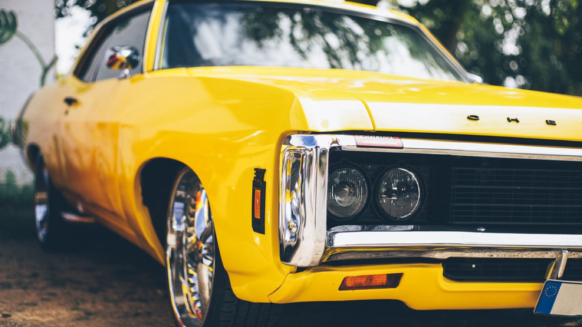 The front quarter of a yellow American muscle car parked in dappled shade.